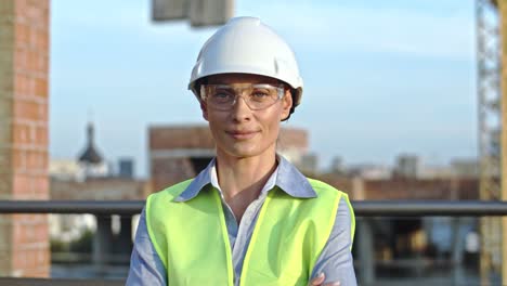 Close-up-of-the-young-pretty-Caucasian-woman-constructor-in-hardhat-and-goggles-looking-straight-to-the-camera-and-smiling-happily-outdoors-at-the-building-site.-Portrait.